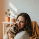 Close-up portrait of a smiling woman in her late 30s holding a small, happy dog. Background is blurred with warm amber and te